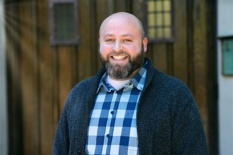 Photo of a smiling man with a brown and gray beard. He is wearing a black sweater and blue and white checked dress shirt. 