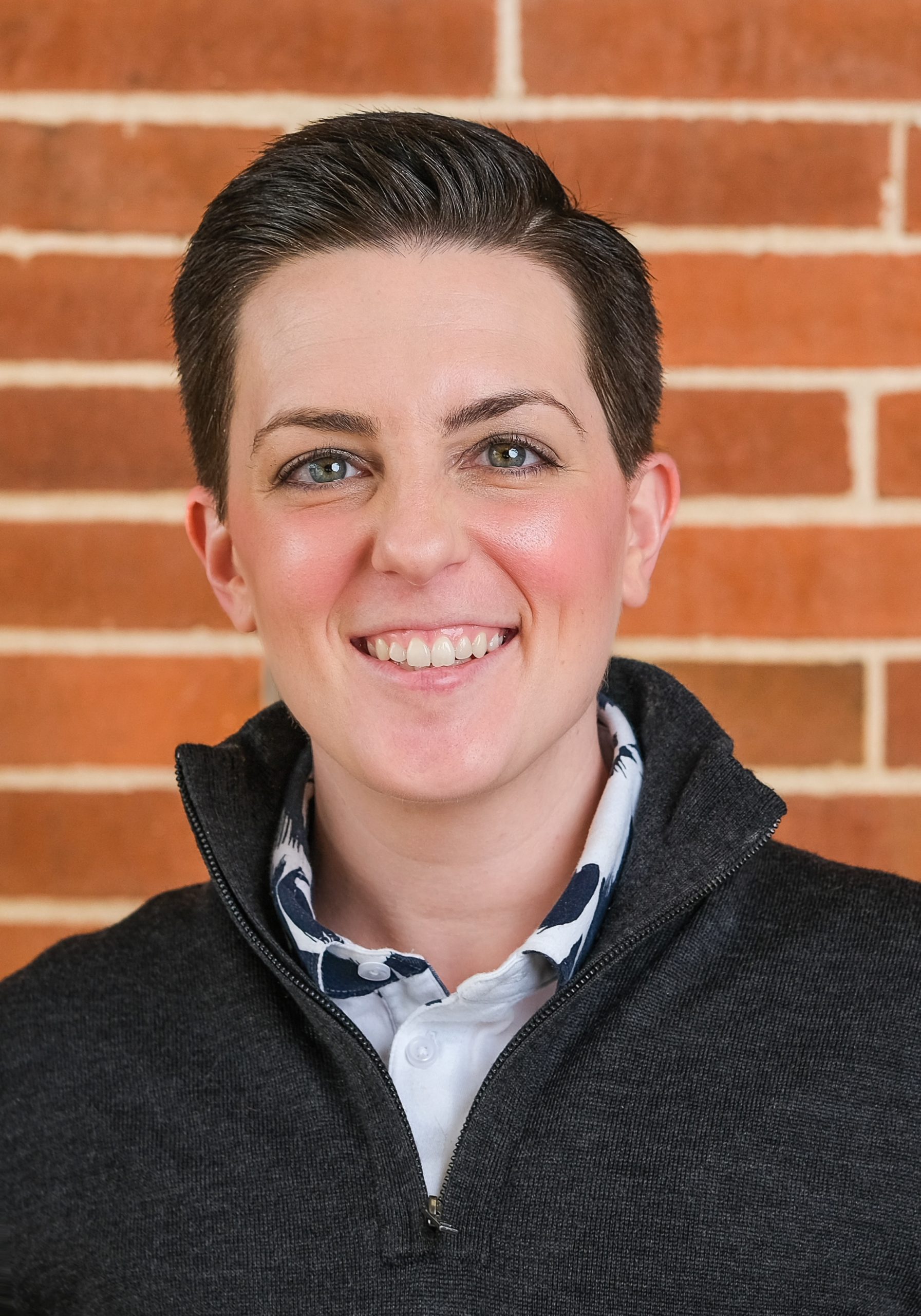 A picture of a woman smiling with short dark hair and a dark jacket in front of a brick wall.