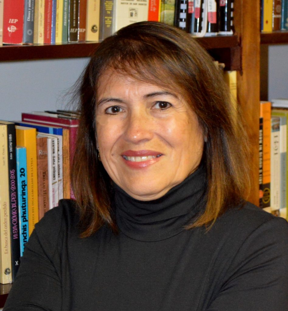 A picture of a woman with long brown hair in a black turtleneck standing in front of some books. 