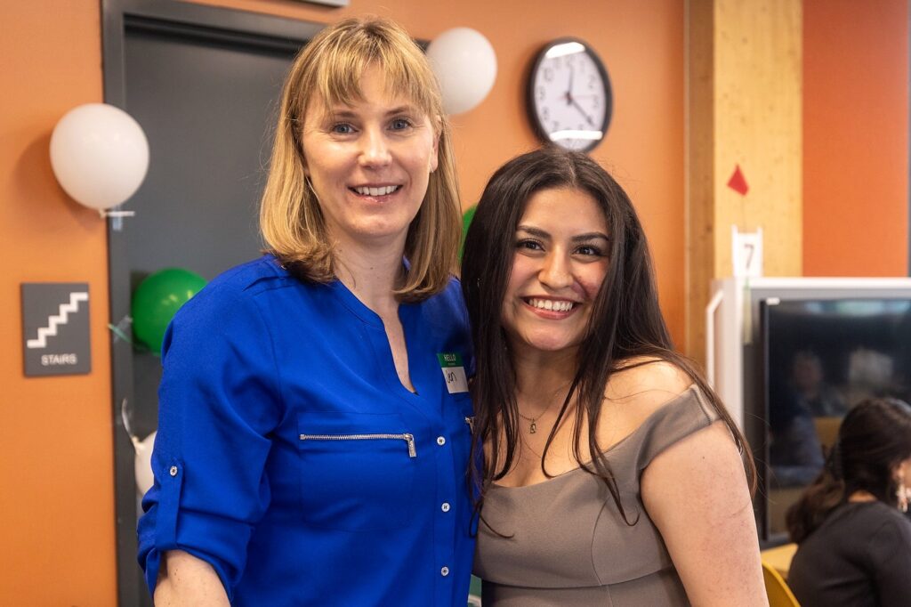 Two women stand smiling together indoors, one wearing a royal blue blouse with a name tag that reads "Jen" and the other in an off-the-shoulder gray dress. The background includes orange walls, a visible clock, and white and green balloons.