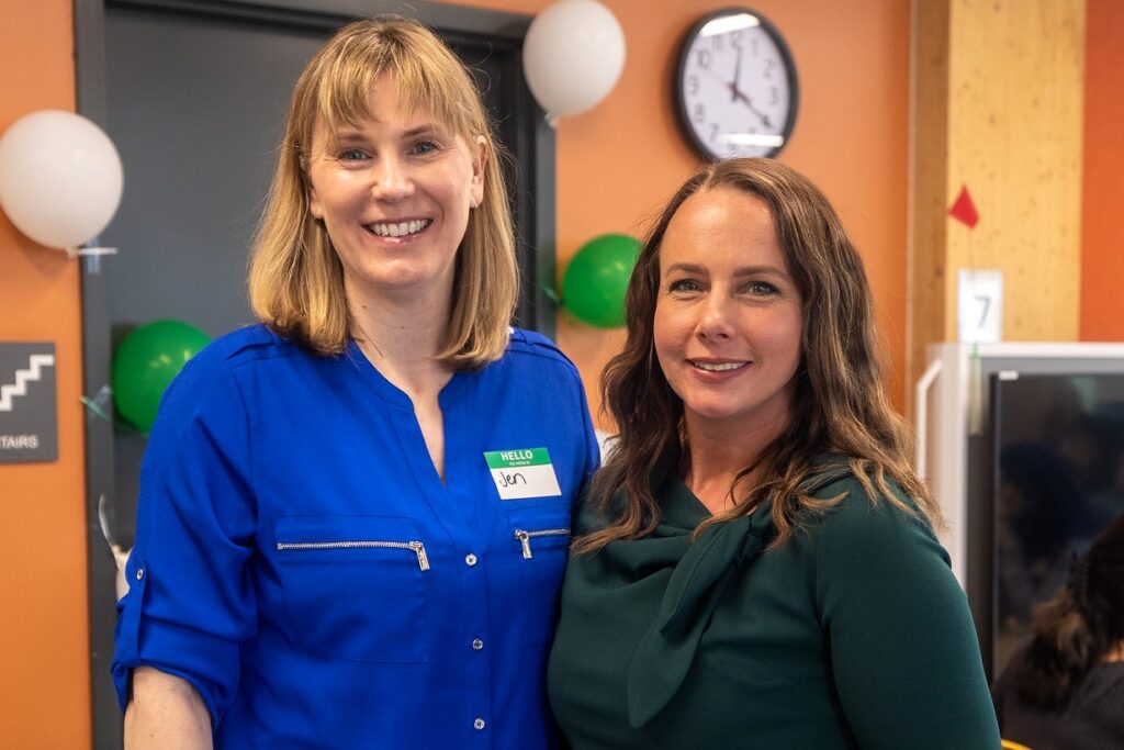 Two women stand smiling together indoors, one wearing a royal blue blouse with a name tag that reads "Jen" and the other wears a dark green blouse. The background includes orange walls, a visible clock, and white and green balloons.