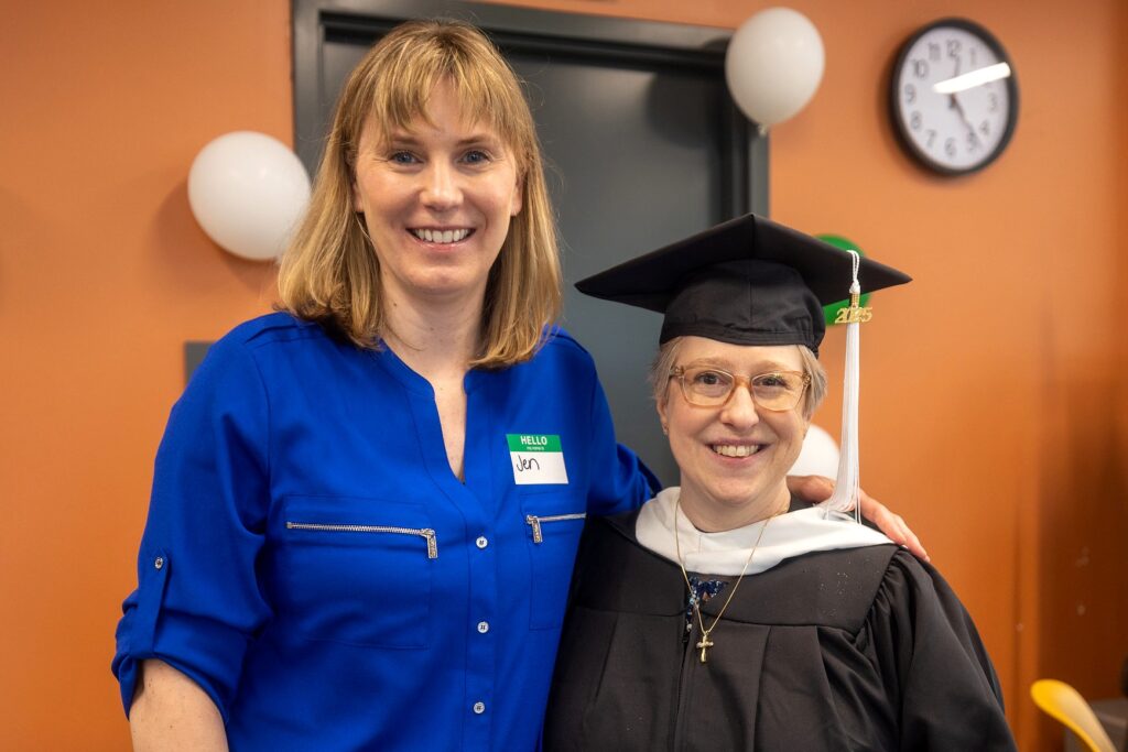 Two women stand smiling together indoors, one wearing a royal blue blouse with a name tag that reads "Jen" and the other woman wears graduation regalia, including a black cap with a "2025" tassel and a gown with a white hood.. The background includes orange walls, a visible clock, and white and green balloons.