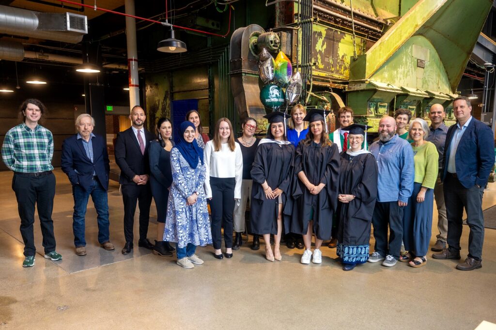 A diverse group of people, including graduates in black caps and gowns, poses for a celebratory photo indoors in front of a large industrial green machine.