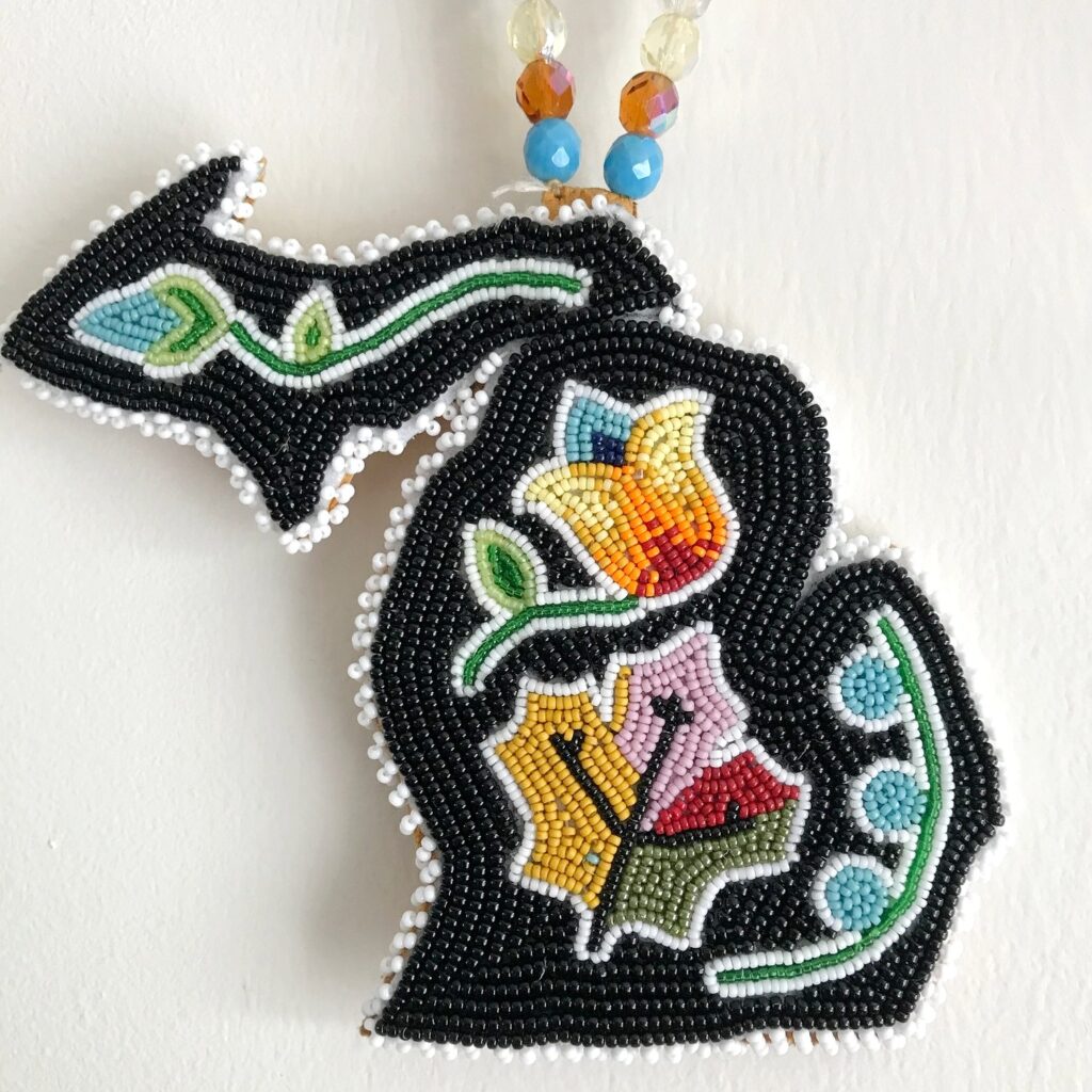 A beaded pendant in the shape of Michigan’s Lower Peninsulas. The black background is adorned with brightly colored floral designs in yellow, red, blue, and green beads, bordered by a trim of white beads. The pendant hangs from a beaded necklace.