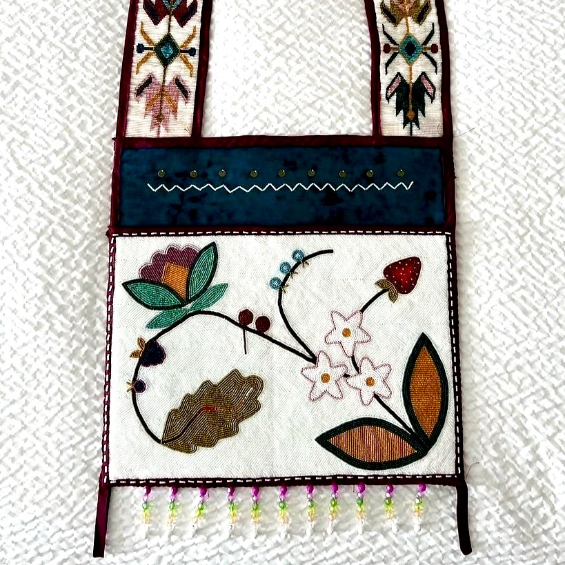 A bandolier bag featuring floral beadwork in vibrant colors on a white background. The strap is decorated with geometric and floral designs, and a row of beaded fringe hangs from the bottom edge of the bag.