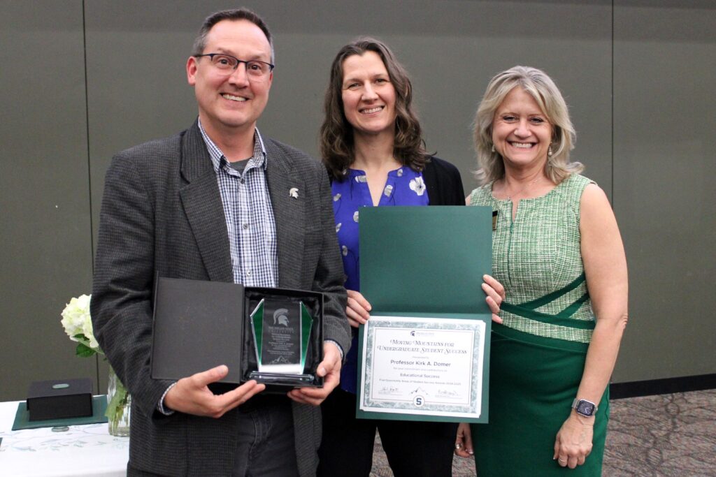 Three people stand together at an awards event. The man on the left is wearing glasses, a gray checked blazer, and a patterned button-down shirt. He holds an open award box displaying a glass plaque with green accents and a Spartan helmet logo. The woman in the center wears a royal blue blouse with a white floral pattern and a black cardigan. The woman on the right is dressed in a green dress with a plaid pattern and a dark green waistband. She holds a certificate awarded to Professor Kirk A. Domer.
