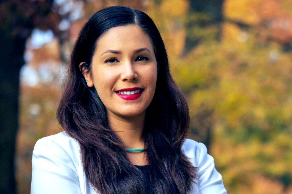 A close-up portrait of a woman standing outdoors in autumn. She has long dark hair, red lipstick, and is wearing a white blazer over a black top with a turquoise necklace. The background shows trees with golden fall foliage.