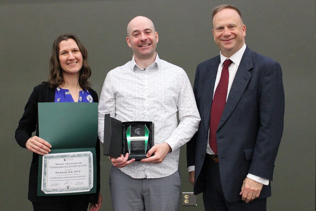 Three people stand side by side in front of a gray wall. The woman on the left wears a royal blue floral blouse and black cardigan and holds a certificate awarded to Pat Arnold. The man in the center wears a white shirt with a small gray pattern and gray slacks, and he holds an open award box. The man on the right wears a navy blue suit, a white dress shirt, and a red tie.