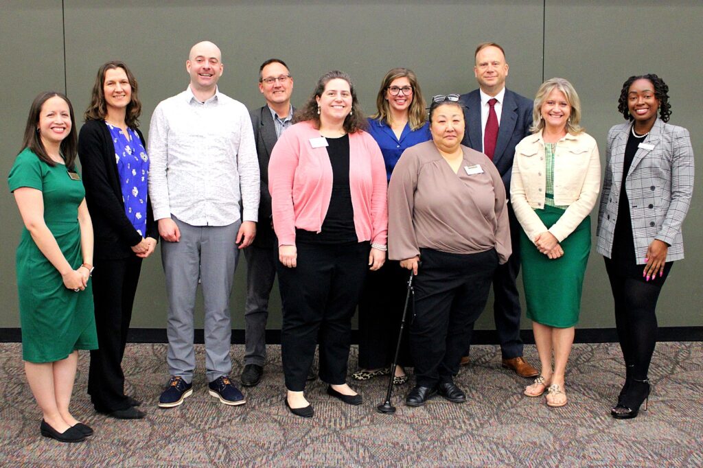 Ten people stand together for a group photo. From left to right: a woman in a green dress with short sleeves; a woman in a royal blue floral blouse with a black cardigan; a man in a white patterned shirt and gray slacks; a man in a gray blazer and checkered shirt; a woman in a black top and pink cardigan; a woman in a bright blue blouse and glasses; a woman in a taupe blouse using a cane; a man in a dark suit and red tie; a woman in a green plaid dress with a cream-colored jacket; and a woman in a black top with a black-and-white checkered blazer. All are smiling and standing in front of a grey wall.