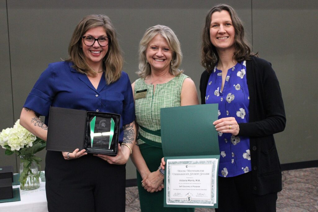 Three women pose with awards. The woman on the left wears a bright blue blouse with rolled sleeves and black pants, and holds an open award box. The woman in the center wears a green plaid dress with a dark green waistband. The woman on the right wears a royal blue blouse with a white floral pattern and a black cardigan, and holds a certificate awarded to Victoria Morris, M.A., for “Self-Discovery of Purpose."