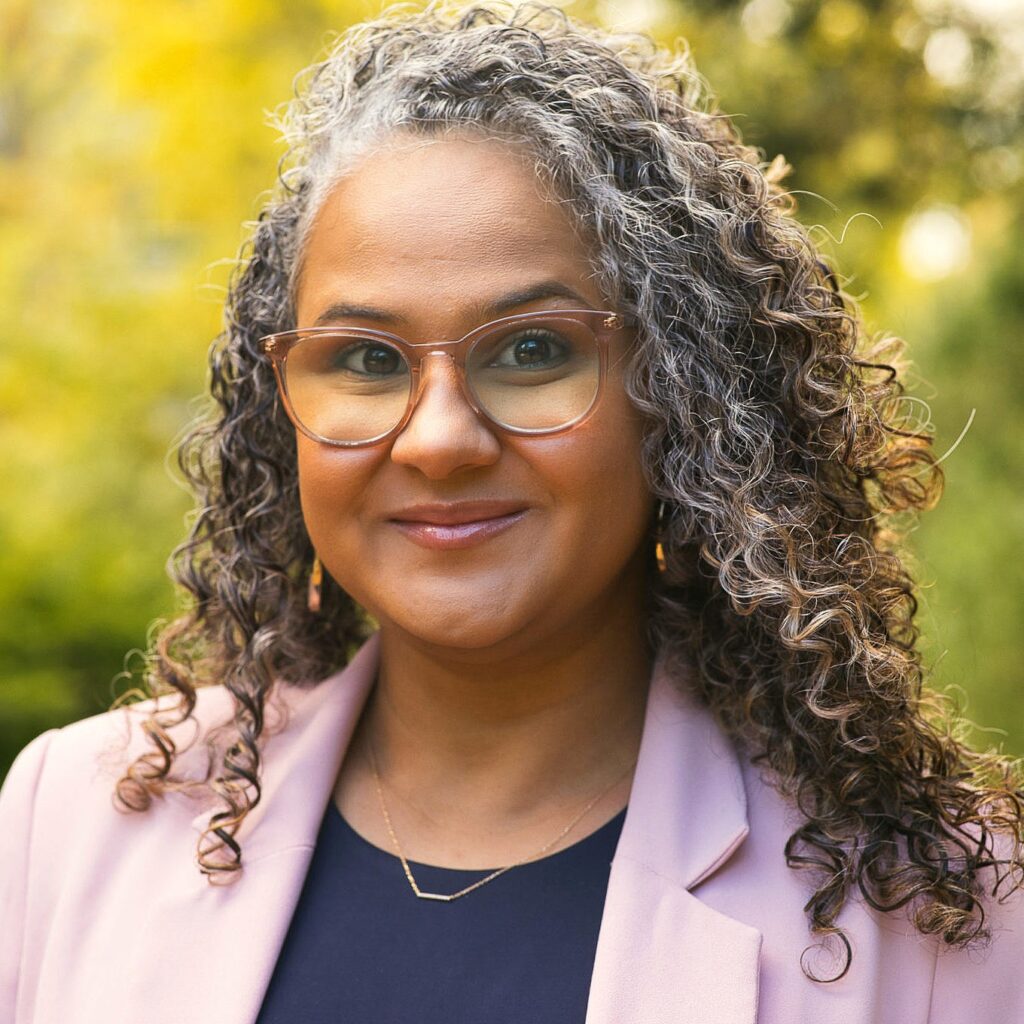 A woman with curly salt-and-pepper hair and clear-framed glasses smiles slightly while standing outdoors with a background of green and yellow foliage. She wears a navy blue top, a light pink blazer, small gold hoop earrings, and a delicate gold necklace.