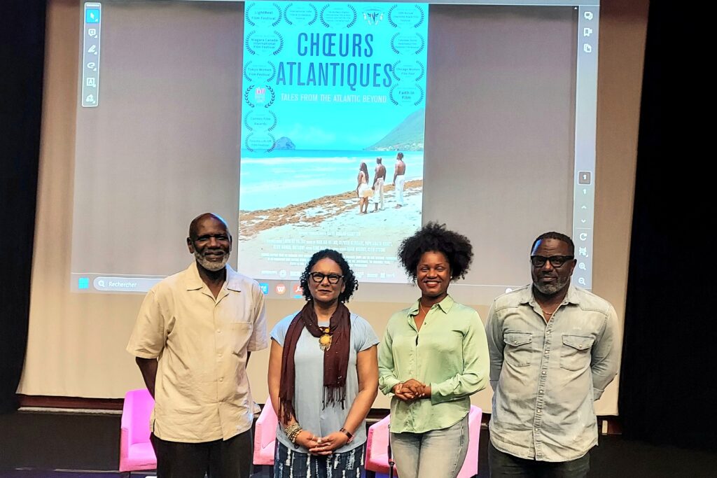 A group of four Black people standing in a row with a movie screen in the background with an image of the “Chœurs Atlantiques” poster projected on the screen.