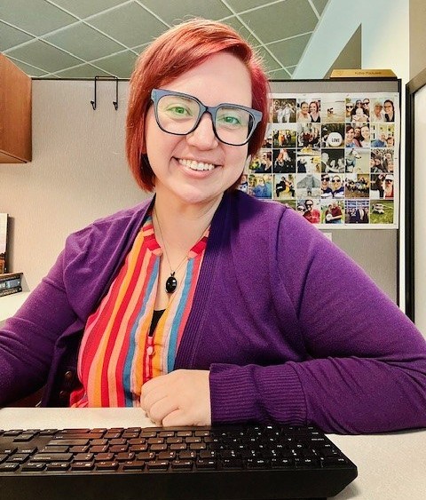 Woman sitting at a desk with a black computer keyboard in front of her. She is wearing a purple cardigan sweater and a bright multi-colored striped top. She has large black-rimmed glassed and short bright red hair. She is smiling and has a lot of photos in the background.