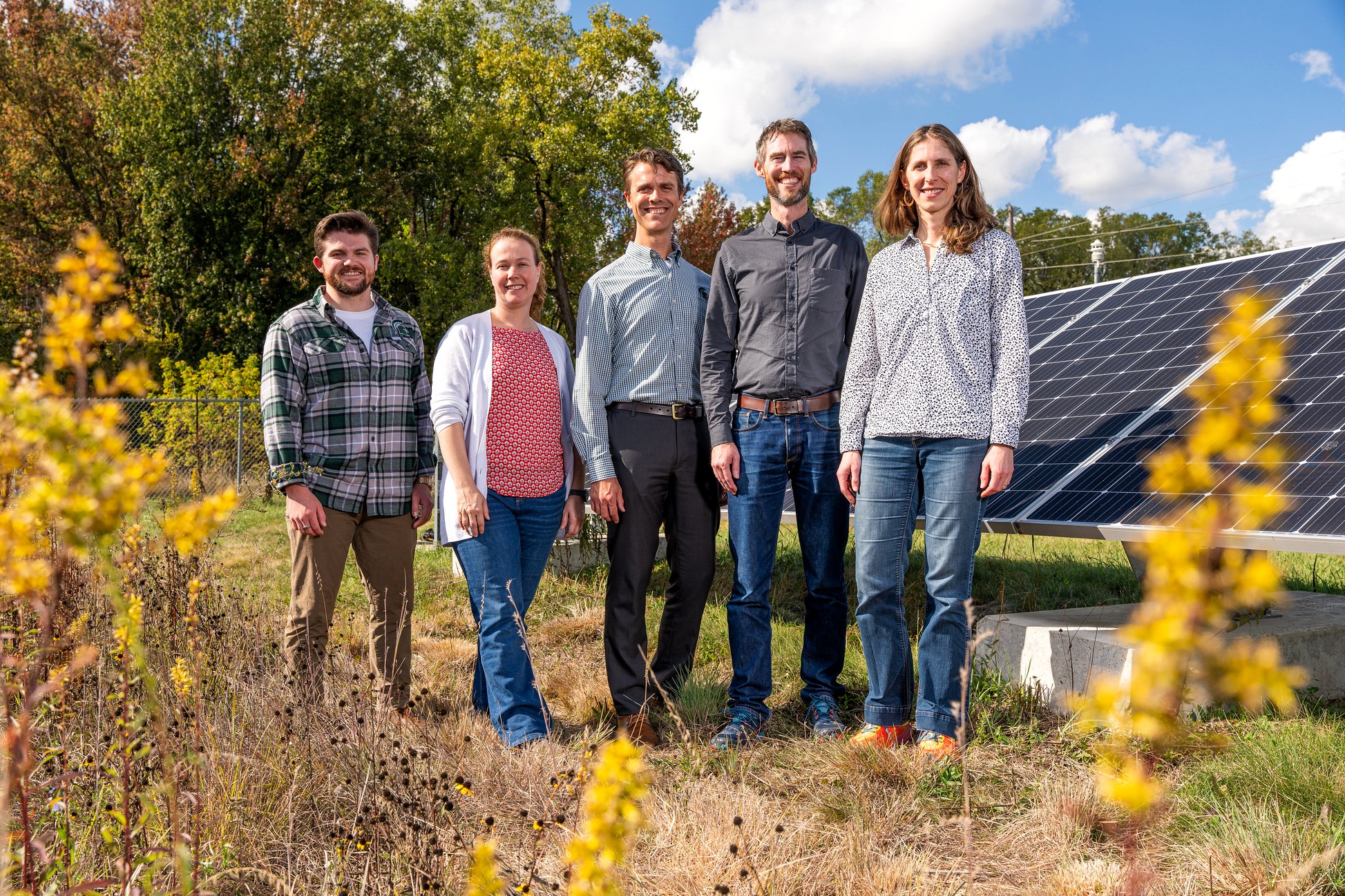 Five people stand together near solar panels in a grassy field with yellow wildflowers in the foreground. Trees and blue sky with scattered clouds are visible behind them.