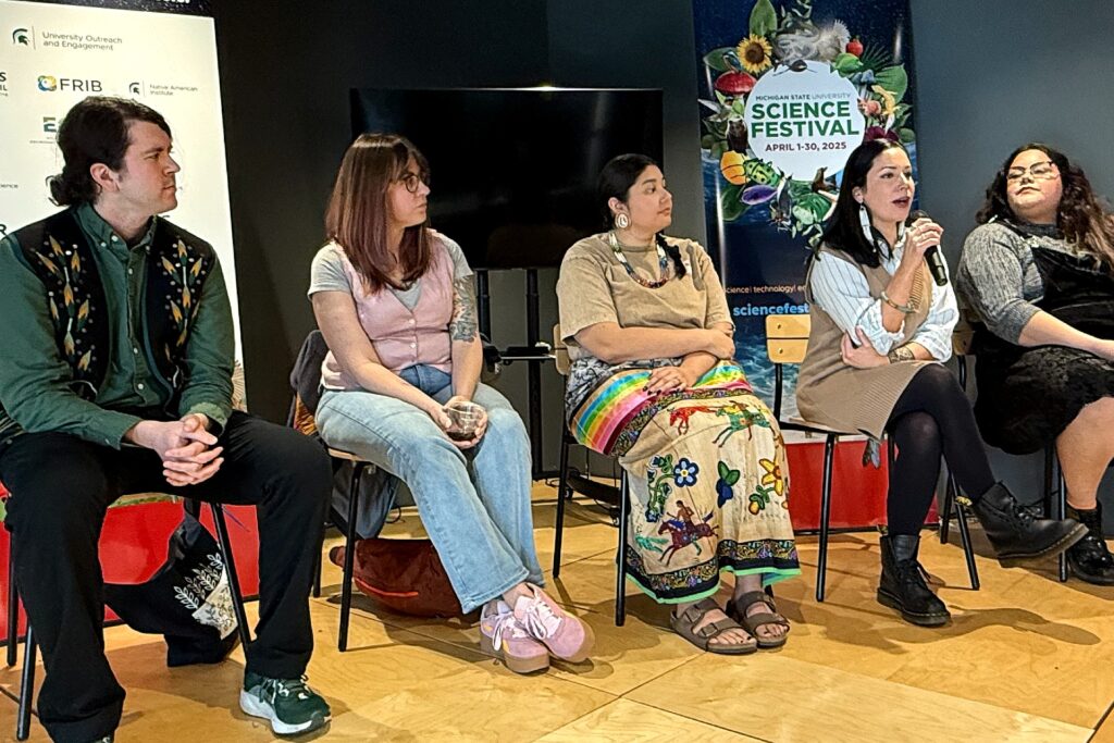 A panel of five speakers seated on a stage at Michigan State University’s Science Festival. One panelist speaks into a microphone while others listen attentively. A banner in the background reads “MSU Science Festival, April 1–30, 2025.”