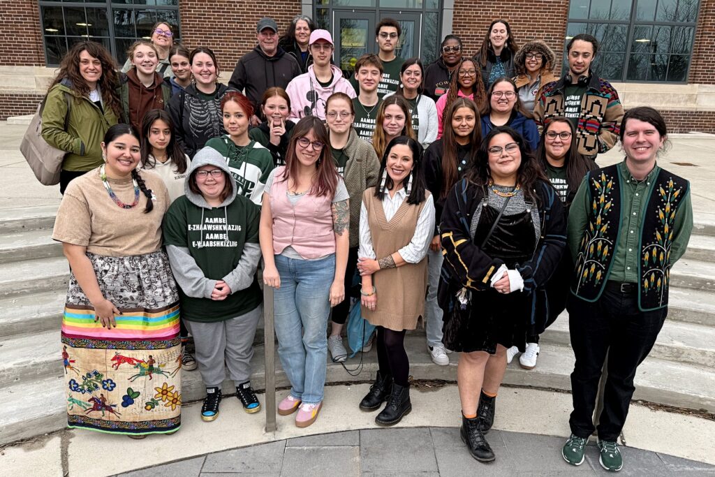 A group of about 30 people gathered on some steps outside. Participants wear MSU apparel and clothing or Indigenous designs. 