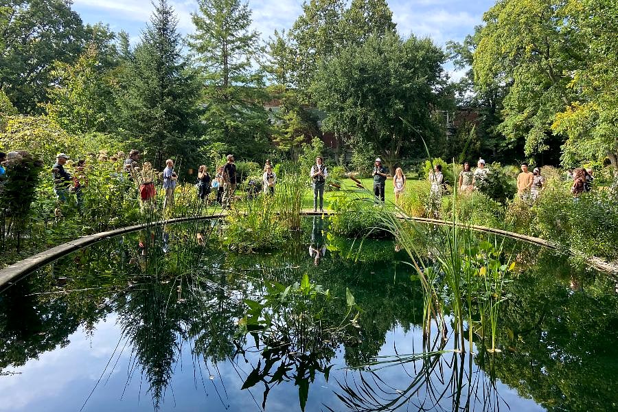 A wide view of the W.J. Beal Botanical Garden shows a group gathered around a circular pond for an outdoor presentation. The speaker stands near the water’s edge while participants listen under a clear sky.