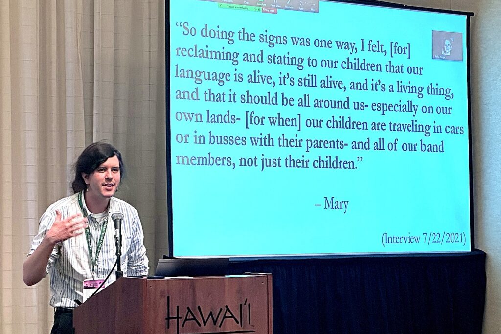 A person presents at a conference podium with a microphone, standing beside a large projection screen that displays a quote about reclaiming and revitalizing Indigenous languages for future generations. 