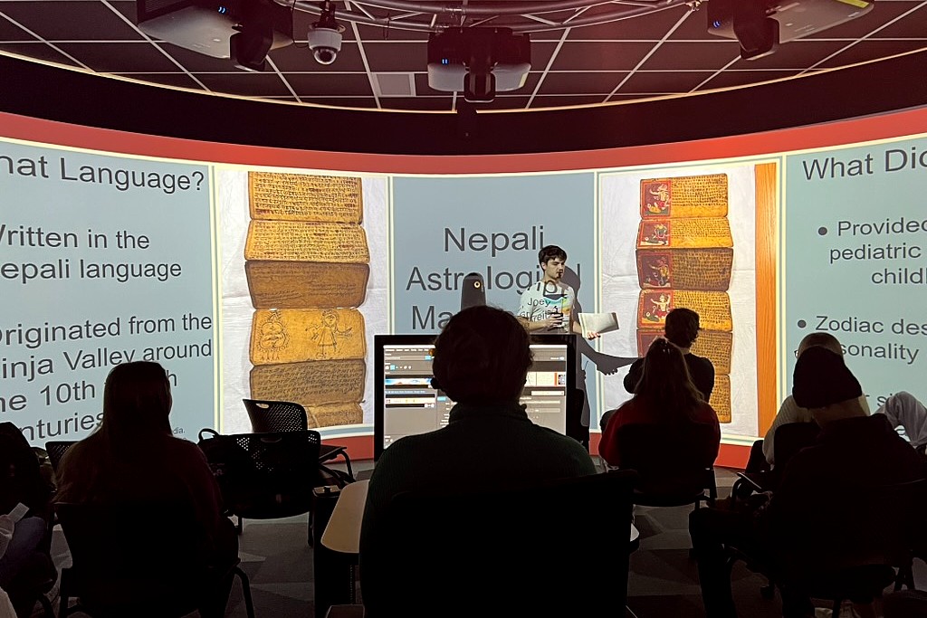 Students sit facing a curved projection screen in a classroom as a student presenter stands at the front discussing Nepali astrology manuscripts. Slides display images of ancient manuscript pages and text about language and historical context, with a computer workstation visible in the foreground.