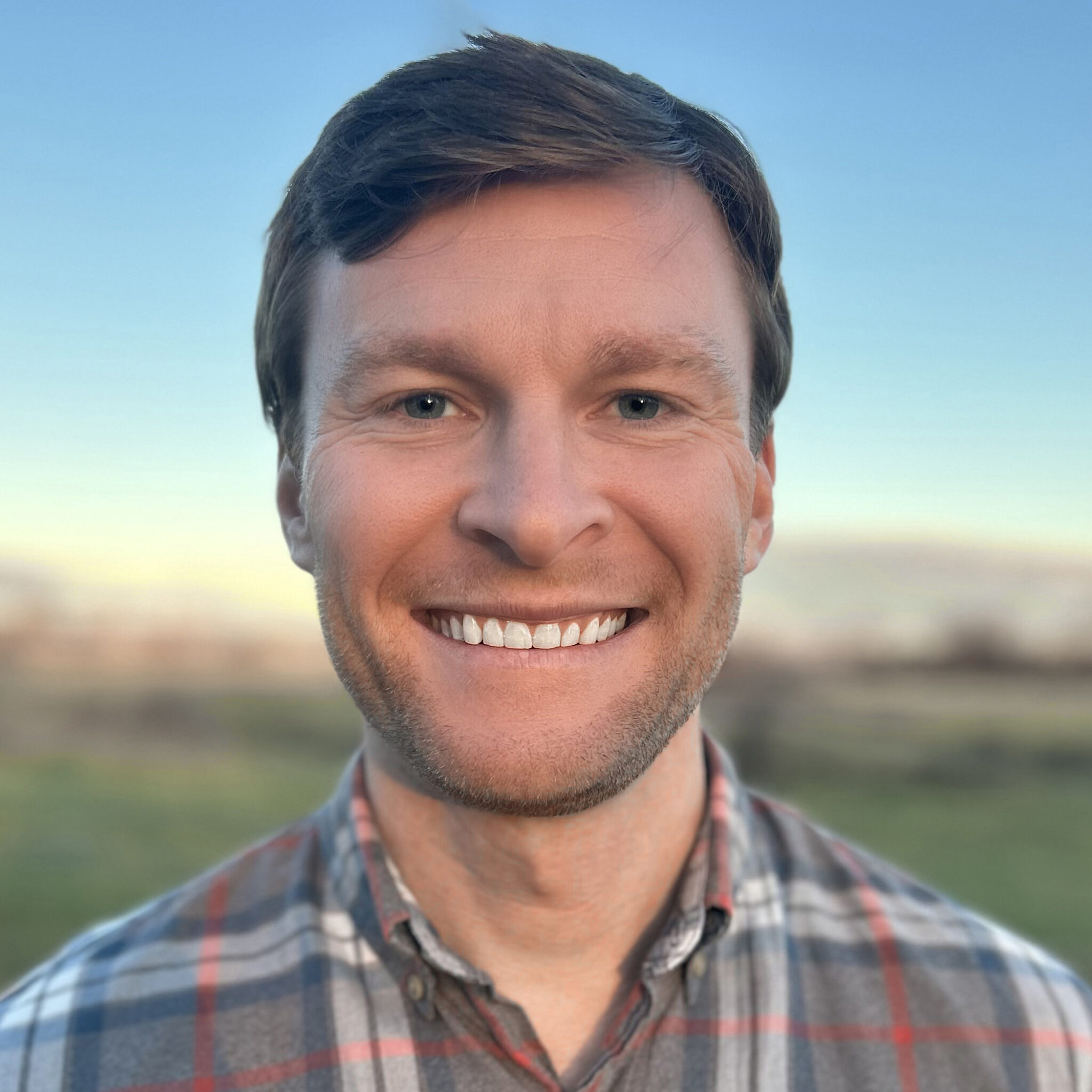 Professional headshot of Joseph Darda smiling against a soft, outdoor background at sunset. He is wearing a gray, red, and blue plaid button-down shirt.