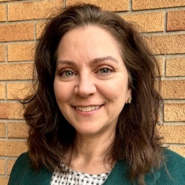 Professional headshot of Laura Ramm with shoulder-length brown hair, wearing a teal jacket over a black and white checkered top. She is smiling in front of a light brown brick wall.