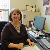 Photo of Joyce Meier seated at a desk in an office setting, wearing a patterned blue and white blouse under a dark purple cardigan. A computer monitor and office supplies are visible in the background.