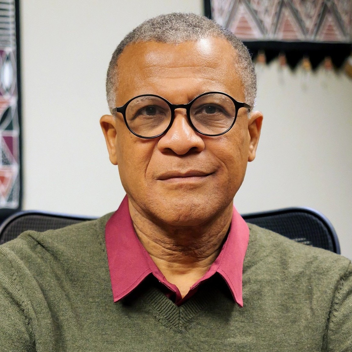 Professional headshot of Philip Effiong wearing round, black-rimmed glasses, a maroon collared shirt, and an olive green V-neck sweater. He is smiling slightly in an indoor office setting.