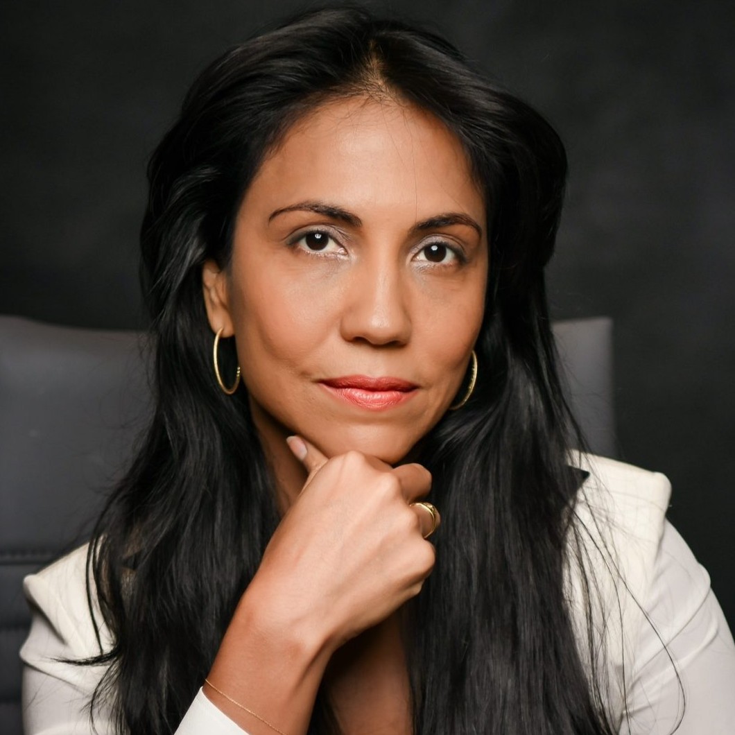 Professional headshot of Elena Ruiz with long, dark wavy hair and gold hoop earrings. She is wearing a white long-sleeved blouse and is posed with her hand resting under her chin against a dark, neutral background.