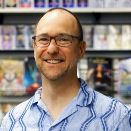 Professional headshot of Ryan Claytor wearing dark-rimmed glasses and a light blue short-sleeved button-down shirt with a dark blue geometric zig-zag pattern. He is smiling in front of a blurred background featuring shelves of comic books.