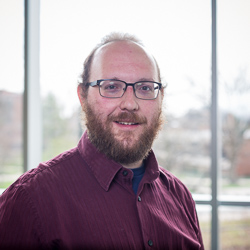 Professional headshot of Aaron Ohlrogge wearing glasses and a maroon button-down shirt. He is smiling in an indoor setting with a large window in the background.