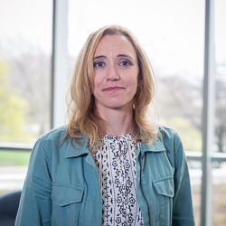 Professional headshot of Amy Kroesche wearing a teal jacket over a white and navy patterned blouse. She is standing in front of a large window with soft outdoor light.