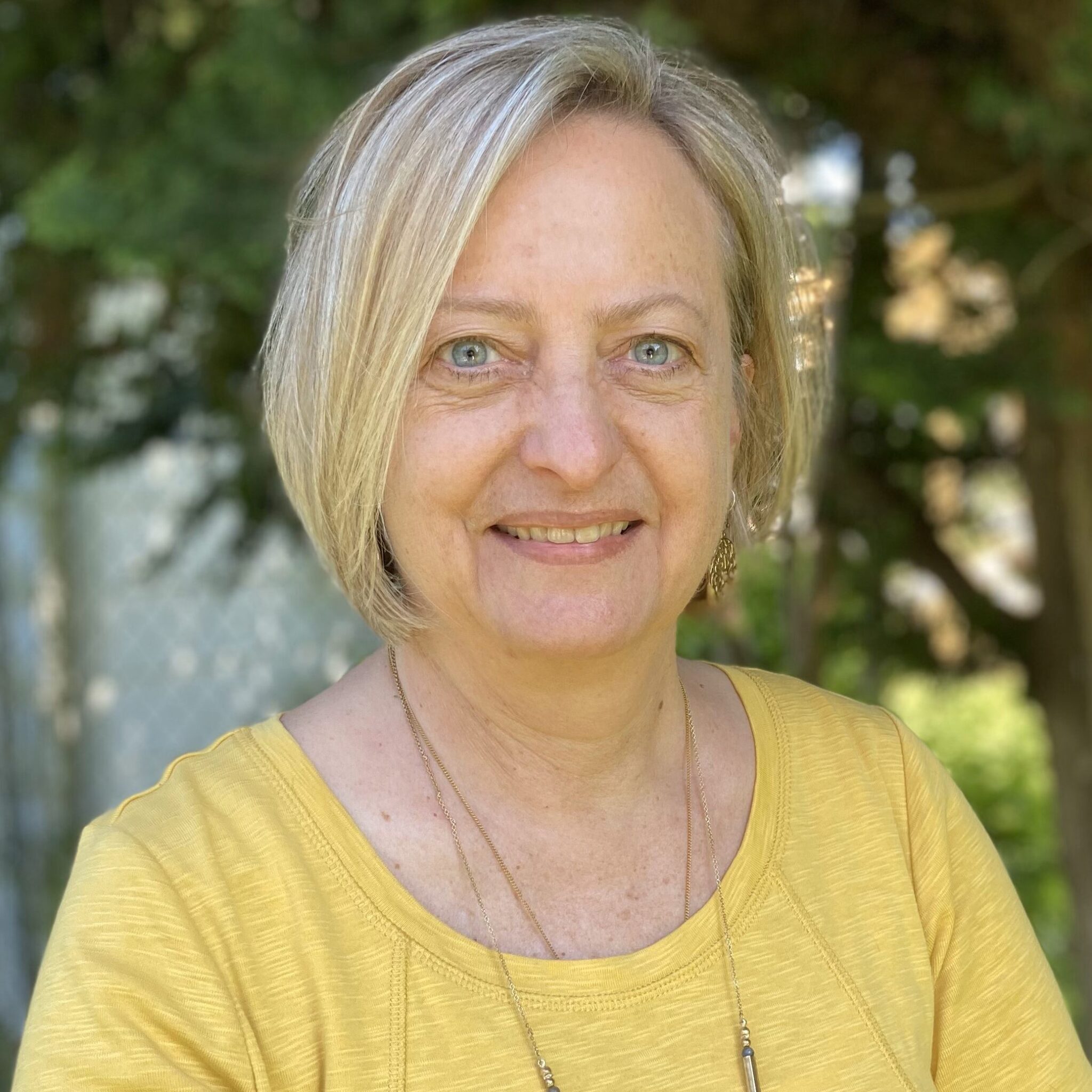 Professional headshot of Laura Smith with blonde hair and blue eyes, wearing a yellow textured shirt and two thin gold necklaces. She is smiling in front of blurred green foliage.