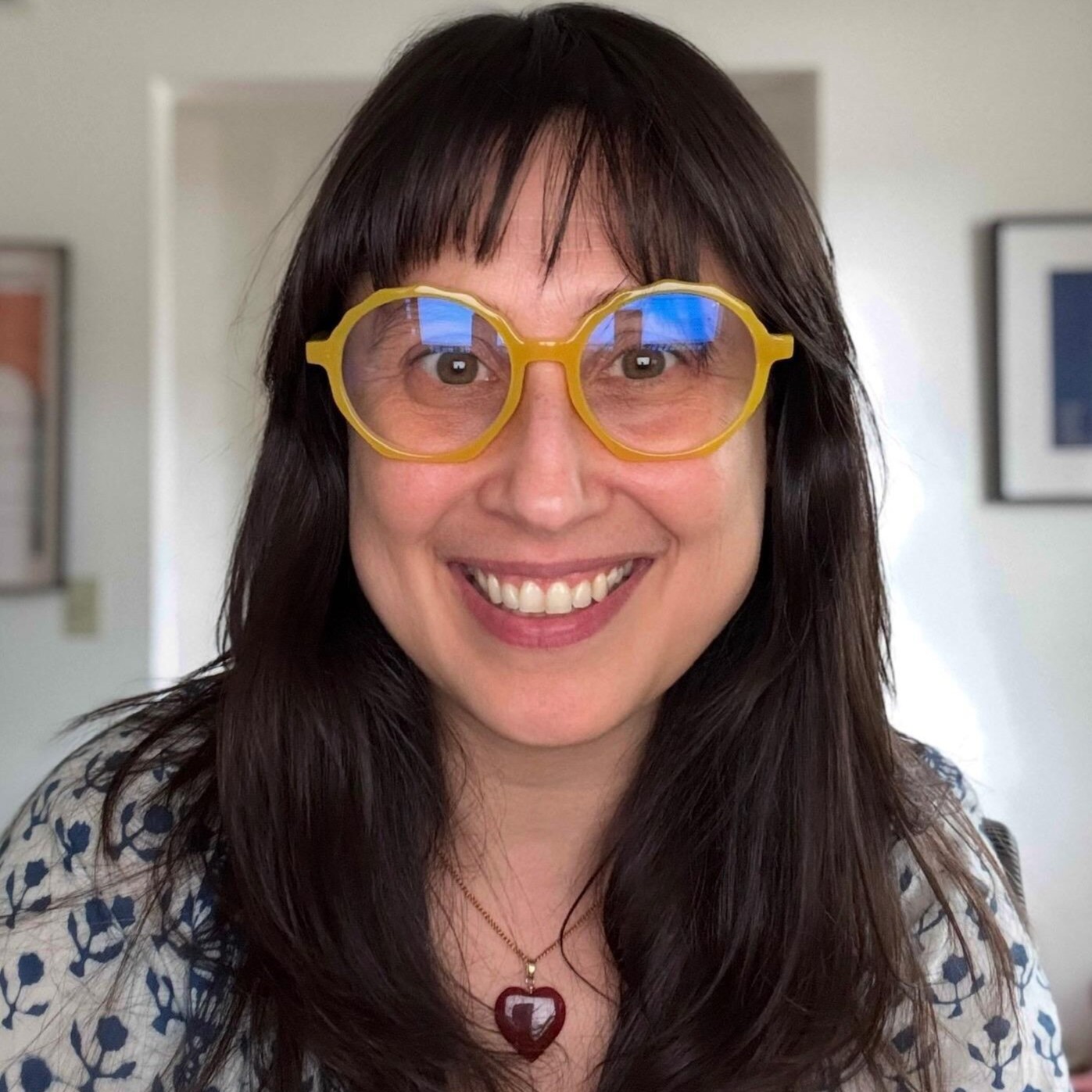 Headshot of Silvina Bongiovanni with shoulder-length dark hair and blue hoop earrings. She is wearing a white boat-neck top with gold button accents on the shoulder and is smiling against a warm, yellow-toned background.