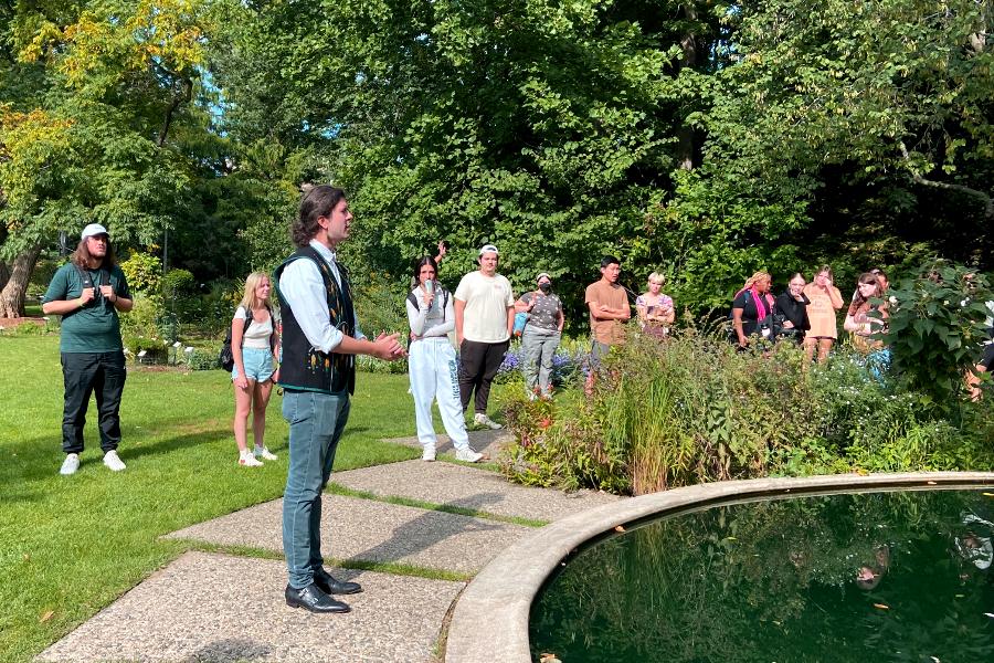 Photo of a person standing next to a small fountain, talking to a group of people in a garden.