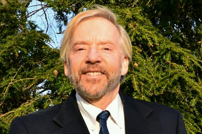 Headshot photo of a man with blonde hair and a beard, smiling, and wearing a black suit and tie and a white button-up shirt. 