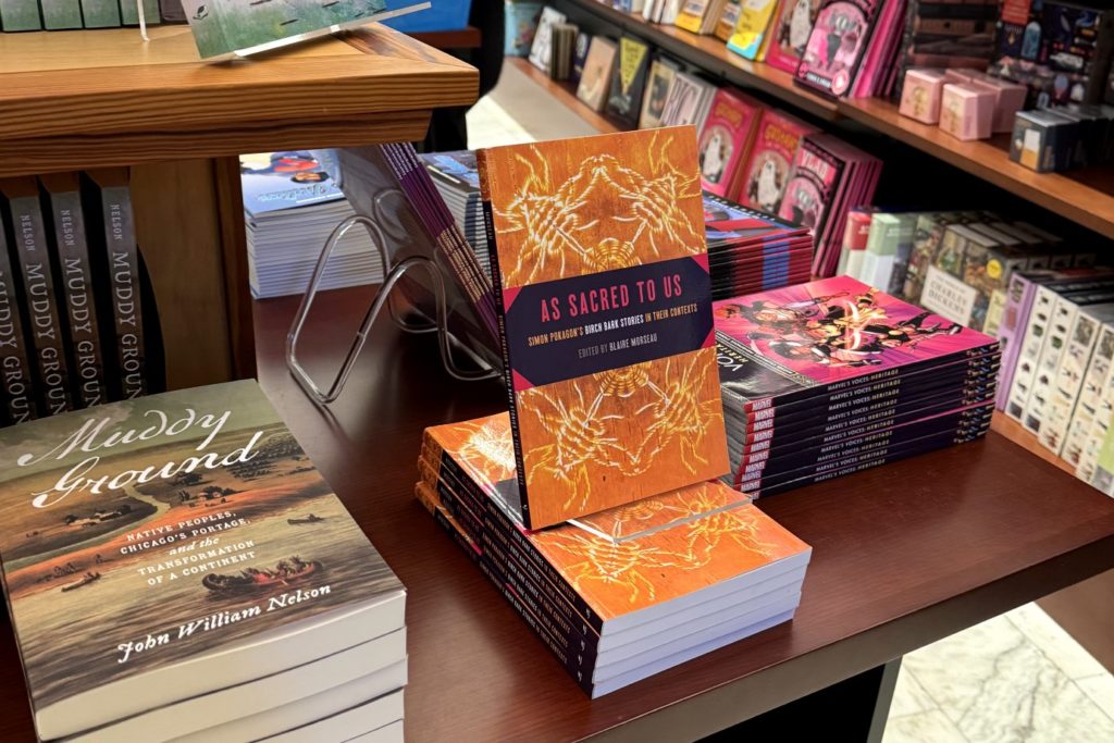 Books on a table and on a shelf in a bookstore with the main book in the photos here having the title of s Sacred to Us: Simon Pokagon' Birch Bark Stories in their Context. 