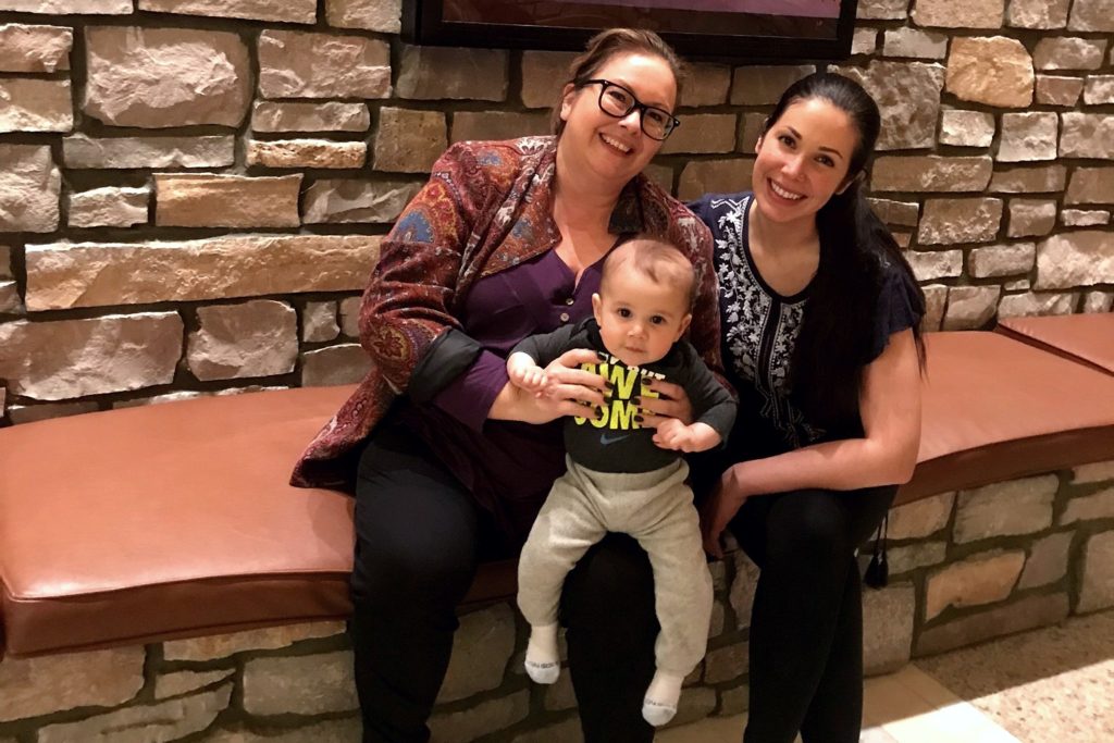 A picture of two women are seated on a leather bench in front of a stone wall, smiling at the camera. The woman on the left is wearing glasses, a purple top, and a patterned jacket, holding a baby in her lap. The baby is wearing a gray shirt with neon green text and light gray pants. The woman on the right is wearing a navy blue embroidered blouse.