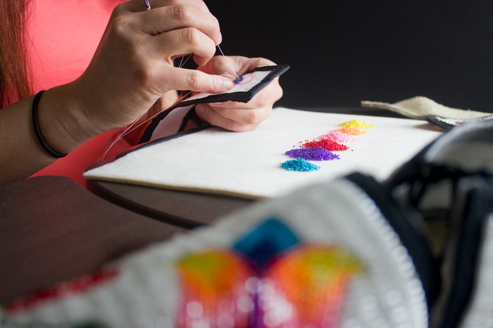 A picture of a pair of hands sewing beads onto fabric, with a row of small, colorful beads arranged on a white mat. Part of a beaded design is visible in the foreground, slightly out of focus.