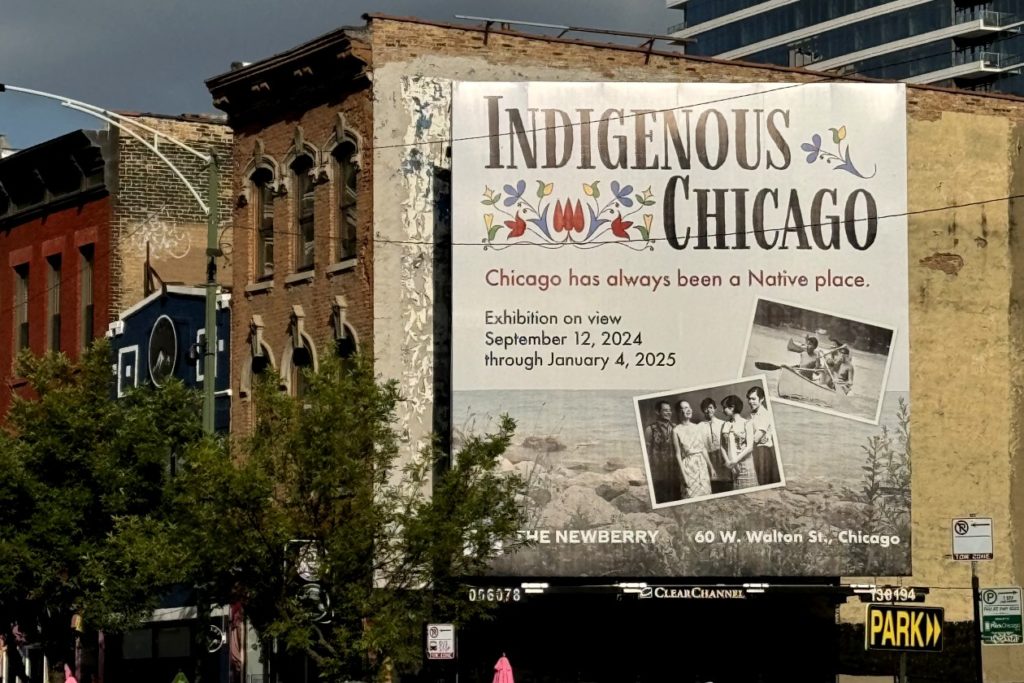 A picture of a large billboard on the side of a brick building reads, "Indigenous Chicago: Chicago has always been a Native place." The billboard includes floral designs and two black-and-white photographs. Trees and other buildings are visible in the foreground and background.