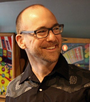 A picture of a man with glasses and black button down posed in front of a pinball machine.